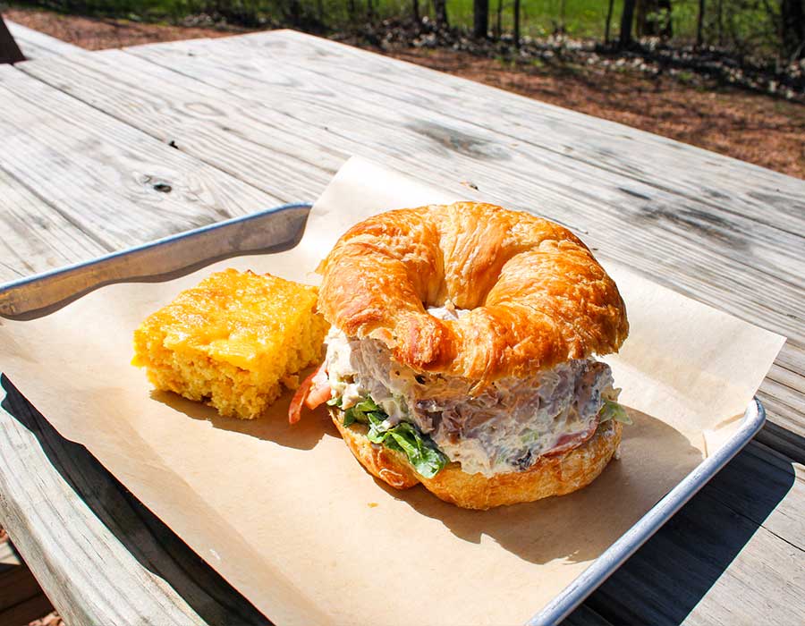 A croissant sandwich and cornbread square from Klinger's Real BBQ Menu on a wooden picnic table.