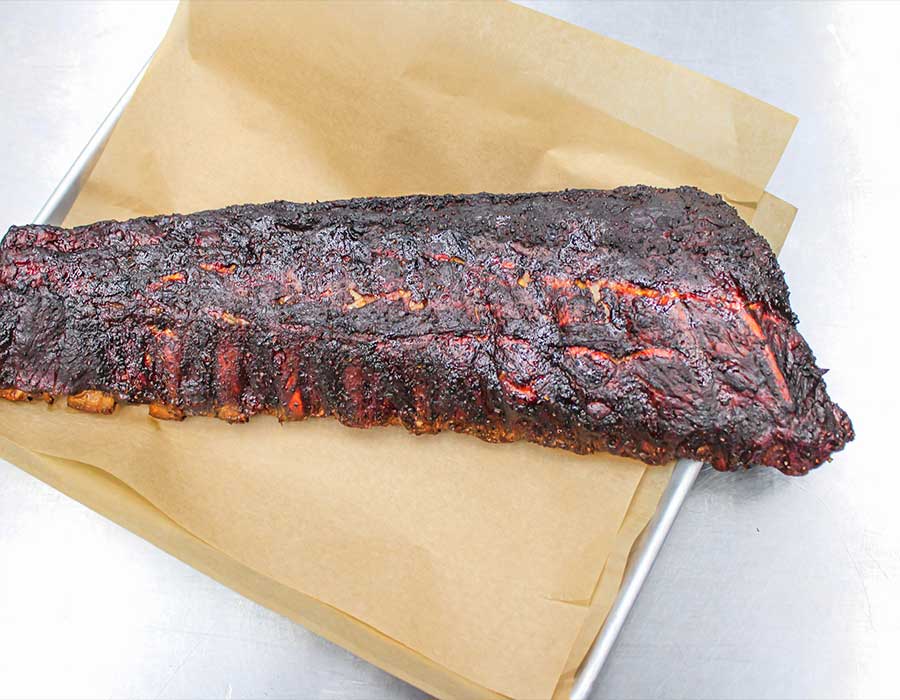 A rack of smoked ribs from Klinger's Real BBQ Menu on parchment in a metal tray.